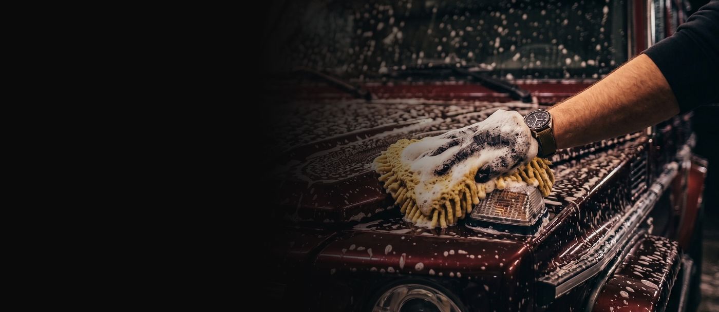 Hand mit eingeschäumtem Waschhandschuh auf der Motorhaube eines dunkelroten Geländewagens